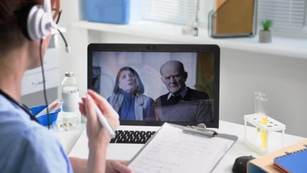 オンライン相談 ヘッドセットを持つ若い女性医師は 医療オフィスのラップトップ上のビデオリンクを介して高齢者男性と女性に助言します — ストック動画