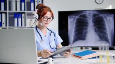 Kulaklık takmış genç bir kadın doktor, tıbbi ofiste otururken hastalarla video bağlantısı yoluyla iletişim kuruyor.