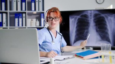 Genç bir kadın tıbbi çalışanın kulaklık kullanarak video konferansında kulaklık kullanarak iletişim kurması, iş arkadaşlarıyla ofiste otururken röntgen sonuçlarını tartışması.
