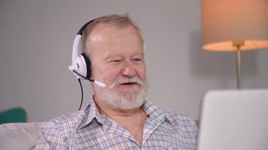 Mikrofon ve kulaklıklı modern gülümseyen yaşlı adam odada otururken video konferansı yoluyla iletişim kuruyor.