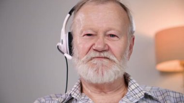 Emeklilik yaşında gülümseyen bir adam mikrofon ve kulaklıkla internetteki video konferansında kameraya bakarak iletişim kuruyor.