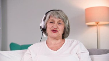 Kulaklık takmış modern yaşlı kadın emekliler rahat bir odada oturup kameraya bakarken web kamerası ve video iletişimiyle iletişim kuruyor.