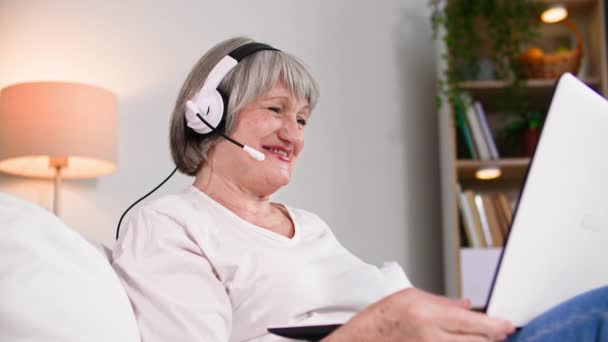 在线交流 微笑的老年女性退休人员在舒适的房间里坐在沙发上 用话筒和耳机通过视频会议与家人交谈 — 图库视频影像
