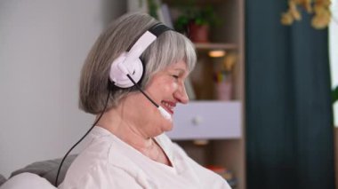 Kulaklıklı yakışıklı yaşlı bir kadın evde dinlenirken video konferansı yoluyla iletişim kurmak için modern teknolojiyi kullanıyor.