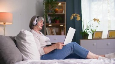 Kulaklıklı neşeli yaşlı bir kadın, yatağında dinlenirken video iletişimi için modern teknolojiyi kullanıyor.