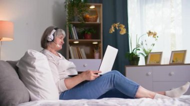 Emeklilik yaşındaki neşeli bir kadın, rahat bir odada yatarken bilgisayar üzerinden iletişim kurmak için kulaklık kullanıyor.