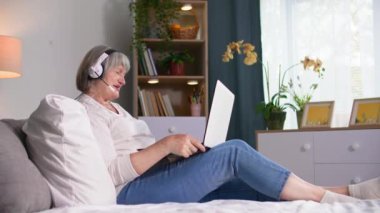 İnternet iletişimi, evde kanepede oturmuş kameraya bakıp gülümseyen, kulaklık takmış yaşlı bir kadın.
