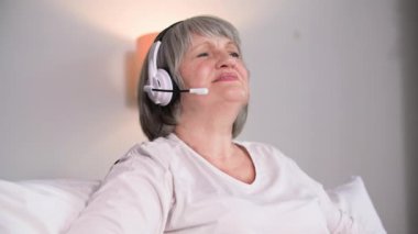 Çekici, olgun bir kadın, odada kanepede otururken bir video konuşmasında kulaklık kullanıyor.