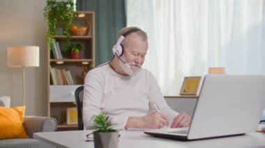 Kulaklıklı ve mikrofonlu yaşlı bir adam laptoptaki bir video görüşmesinde konuşur ve masada otururken not defterine not alır.