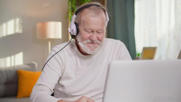一位留着灰胡子的现代男性养恤金领取者坐在舒适的房间里的桌子旁 一边用耳机在笔记本电脑上通过视频通话 一边在记事本上记笔记 — 图库视频影像