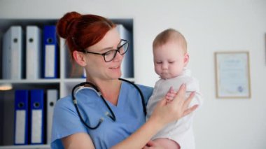 Bir tıp ofisinde kucağında küçük bir çocuğu tutan, gülümseyen ve kameraya bakan bir kadın çocuk doktoru portresi.