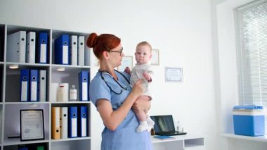 Kadın bir pediyatri doktorunun portresi. Bir muayenehanede kucağında bir bebek var. Gülümsüyor ve kameraya bakıyor.