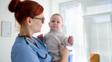 Gözlüklü genç bayan çocuk doktoru, doktordaki hastayı muayene ederken kucağında küçük bir çocuk tutuyor.