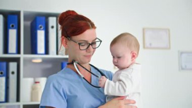 Çocukla ilgilenen, gözlüklü bir kadın doktor ve kollarında küçük bir çocuk hastaları kabul ederken sağlık odasında duruyor.