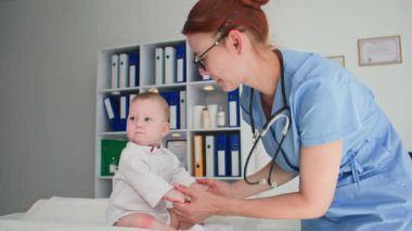 Genç bayan doktor küçük bir bebeği okşuyor ve hastayı muayene ederken onu kollarına alıyor.