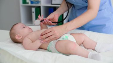 Bebek bezli şirin bir çocuk bebek bezi masasında yatıyor ve doktorunun muayenesi sırasında steteskobu ağzına çekiyor.
