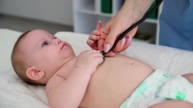 Bebeğe bakan, fonendoskop kullanan bir kadın doktor, muayene odasındaki pelinasyon masasında küçük bir çocuğun nefes alışını dinler.