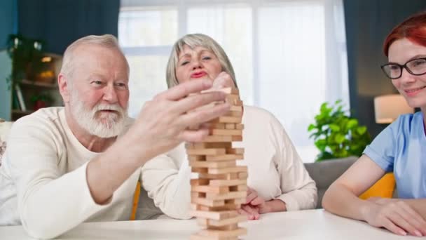 老年人的社会适应 穿着医疗制服的女性社会工作者和一对老夫妇坐在房间的桌子旁玩棋盘游戏 — 图库视频影像