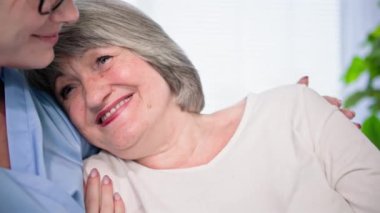Yaşlı, genç, tıbbi üniformalı bir kadın çalışana bakmak, odadaki kanepede oturan yaşlı kadını destekliyor.