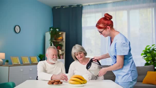 为了帮助一对老年夫妇 一位年轻的女护士在房间里吃点心的同时 照顾着一位老年男女 将热水倒入杯子里 — 图库视频影像