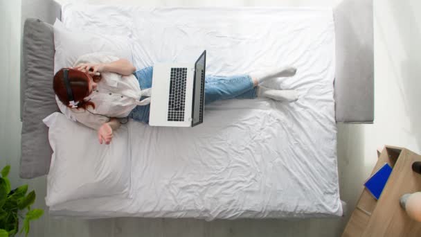 自宅で仕事 女性は友人と通信するためにノートパソコンのヘッドセットを使用したり 部屋のベッドに横たわりながら仕事 トップビュー — ストック動画