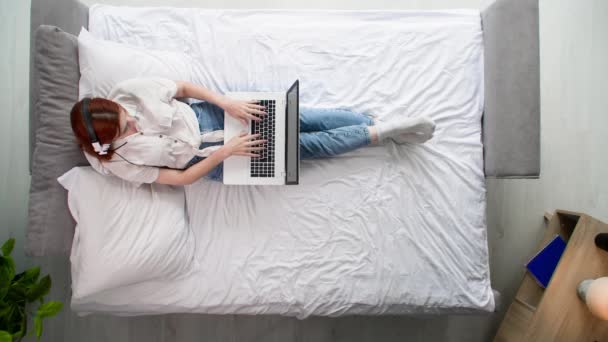 一个自由职业的年轻女人 带着耳机和笔记本电脑 躺在床上 一边打电话一边聊天 一边放眼望去 — 图库视频影像