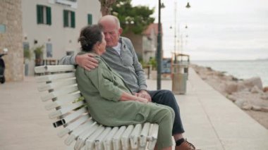 Evli çiftler birlikte bir bankta oturup deniz manzarasının tadını çıkarıyorlar.