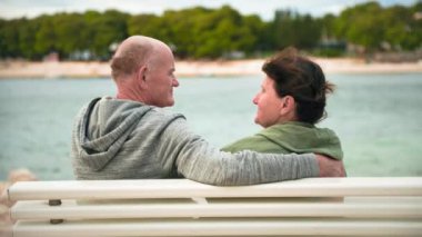 Yaşlı kadın ve erkek turistler, emeklilerin tatili sırasında sıcak bir günde deniz kıyısındaki bir bankta kucaklaşırlar.