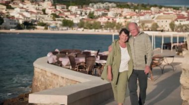 Aktif yaşlılık, neşeli yaşlı adam ve kadın güneşli bir günde ortak emeklilik tatili sırasında deniz kıyısında birlikte yürüyorlar.