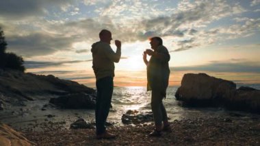 Aktif emekliler, yaşlı adam ve kadın deniz kıyısında güneş batarken suya taş atarken eğleniyorlar.