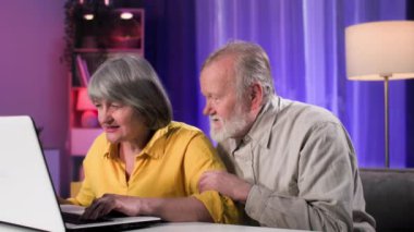 Neşeli yaşlı adam ve kadın rahat bir odada otururken online bir dizüstü bilgisayarla modern bilgisayar oyunları oynayarak eğleniyorlar.