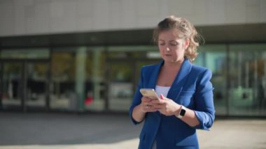 Akıllı telefonlu takım elbiseli genç bir iş kadını güneşli bir günde bir iş merkezinin arka planında yürüyor.