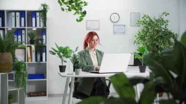 Çevre dostu, gözlüklü genç bir kadın ofiste çiçeklerle otururken dizüstü bilgisayarda çalışıyor.