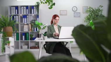 Genç bayan girişimci yeşil bitkilerle dolu bir ofiste dizüstü bilgisayarda çalışıyor.