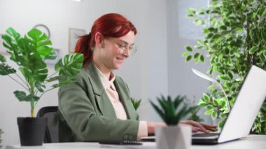 Gözlüklü genç, neşeli bayan iş kadını, yeşil ofisteki masada otururken müşterilerin taleplerini işliyor.