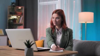 Evde dizüstü bilgisayarla çalışırken not tutan takım elbiseli ve gözlüklü genç bir kadının portresi. Gülümsüyor ve kameraya bakıyor.