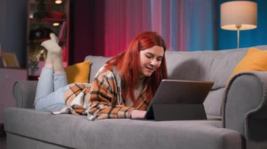 Evde dinleniyor, kaygısız genç bir kadın elinde aletlerle eğleniyor ve akşamları kanepede yatarken video oyunları oynuyor.