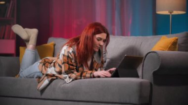 Genç kızıl saçlı kadın arkadaşlarıyla sohbet etmek için tablet kullanıyor ya da akşamları kanepede uzanmış online alışveriş yapıyor.