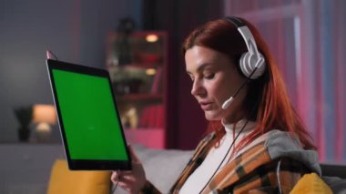 Video çağrısı, kulaklıklı genç bir kadın arkadaşlarıyla iletişim kurmak için modern teknolojiyi kullanıyor. Akşamları kanepede otururken elektronik bir cihaz kullanıyor. Yeşil ekranda.