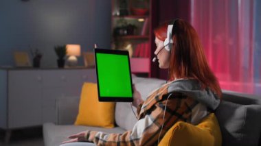 Elinde mikrofon, kulaklık ve tablet olan genç bir kadın şık bir odada kanepede otururken video konuşuyordu, kromakey.