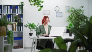 Çevre dostu iç mekan, kadın ofis çalışanı dizüstü bilgisayarda çalışıyor ve yeşil bitkilerin arasında otururken notlar alıyor