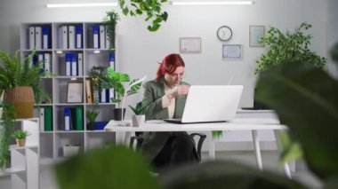 Genç bir kadın yeşil bitkilerle dolu bir ofiste otururken toza ve çiçeklere alerjisi olduğu için laptopta çalışıyor ve hapşırıyor.