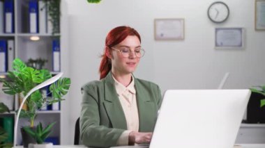 Gözlüklü genç bayan şirket yöneticisi bir dizüstü bilgisayarda çalışıyor ve rahat bir odada otururken notlar alıyor.