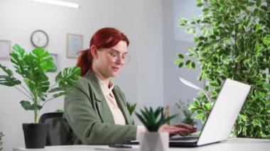 Gözlüklü genç bir kadın dizüstü bilgisayarda çalışıyor. Çevre dostu bir ofiste bitkilerle masada otururken bir not defterine yazıyor.