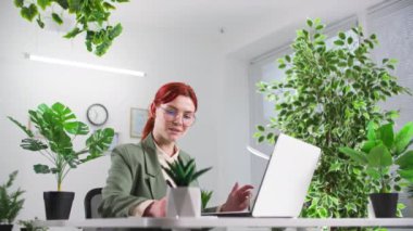 Gözlüklü kadın ofis çalışanı. Odada bitkilerle bilgisayar başında çalışıyor.