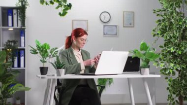 Gözlüklü genç kadın, çevre dostu ofiste çalışırken iş arkadaşlarıyla sohbet etmek için akıllı telefon kullanıyor.