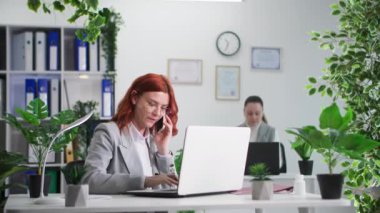 İş günü, meşgul bir kadın telefonlarını cevaplıyor bilgisayarın başında yeşil bitkilerle oturuyor.