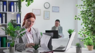 Gözlüklü genç bir iş kadınının portresi dizüstü bilgisayarda çalışırken masada oturan, gülümseyen ve kameraya bakan bir meslektaşı.