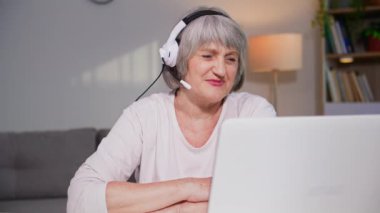 Modern teknoloji, kulaklığı olan çekici olgun bir kadın, rahat bir odada otururken bilgisayarla konuşuyor.