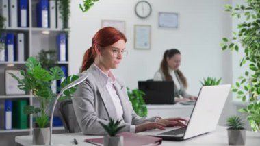 Çalışma ortamı, gözlüklü genç bir kadının portresi dizüstü bilgisayarda çalışırken meslektaşının ofis kağıtlarını fırlatıp kameraya bakması.